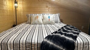 Ein Schlafzimmer einer Holzhütte mit einem Bett, gestreifter Bettwäsche, zwei Kissen mit Tiermotiven, einem Nachttisch, einer Lampe und einer Deckenleuchte.