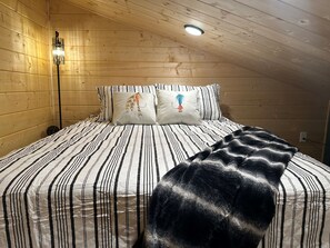 Un dormitori de cabana de fusta amb un llit, roba de llit a ratlles, dos coixins amb dibuixos d'animals, una taula de nit, una làmpada i una llum al sostre.