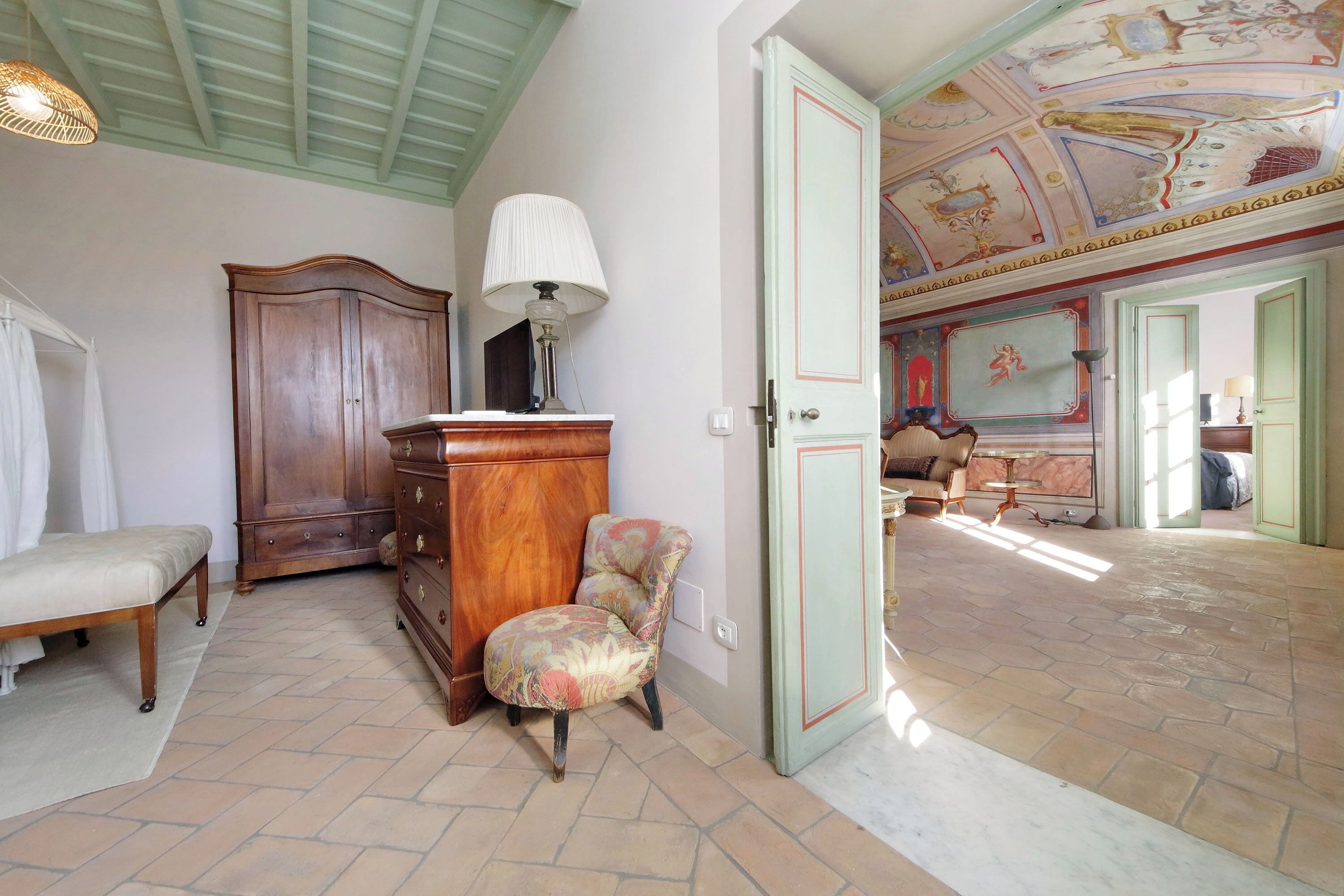 Una habitación con una cama, un armario de madera, una silla y una puerta que da a otra habitación con piso estampado y techo decorado.