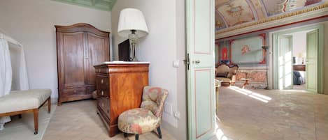 A room with a bed, a wooden cabinet, a chair, and a door leading to another room with a patterned floor and a ceiling with artwork.