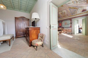 A room with a bed, a wooden cabinet, a chair, and a door leading to another room with a patterned floor and a ceiling with artwork.