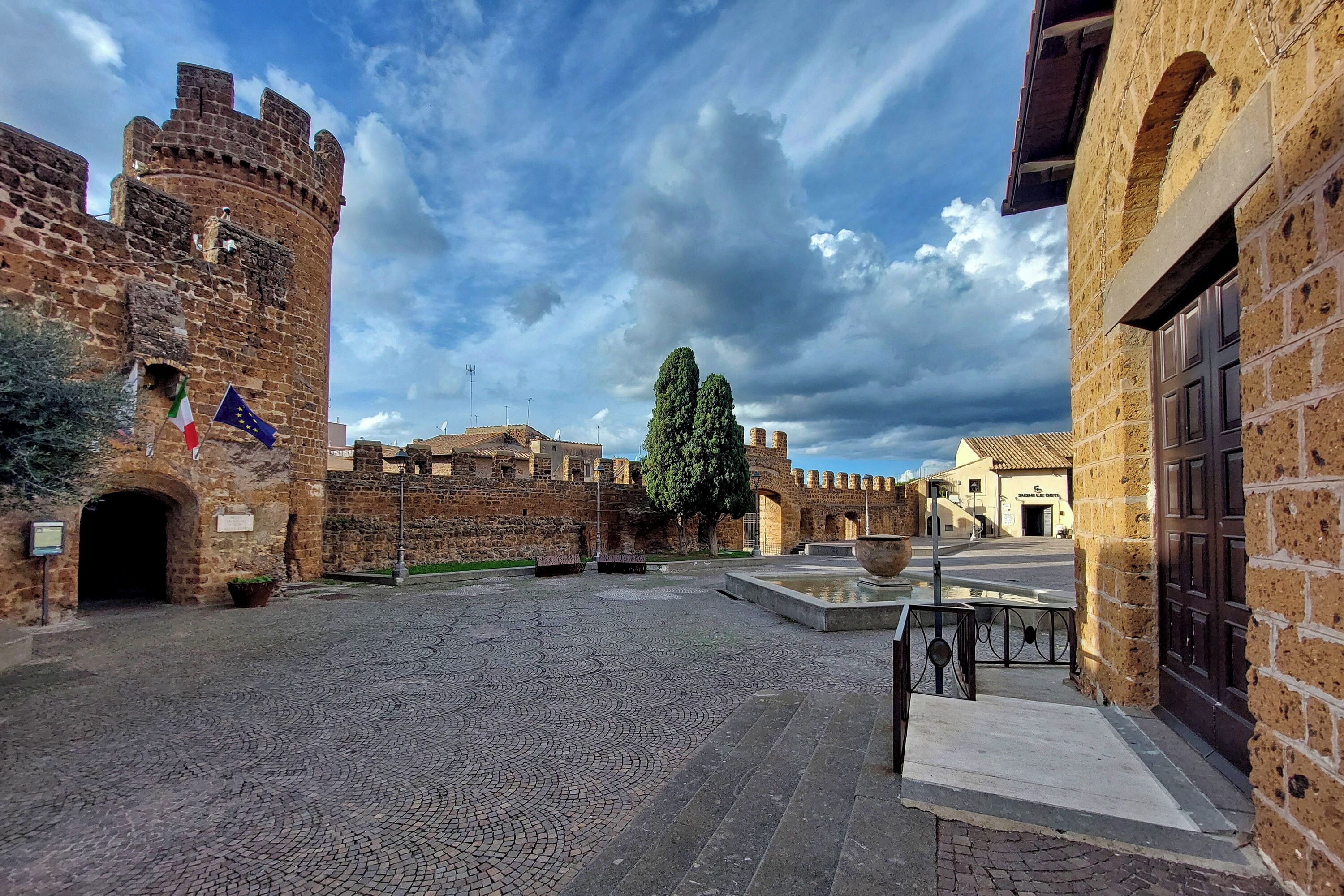Un cortile con pavimentazione in pietra, un albero grande e una struttura simile a un castello, con mura in pietra e una bandiera.