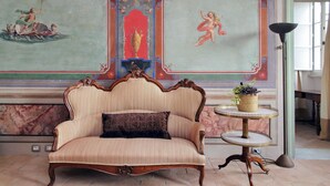 A vintage sofa with a patterned cushion, a small round table with a plant, and a mural with classical figures. - Hotel PalazzoVittoria HeritageCollection (Cerveteri)