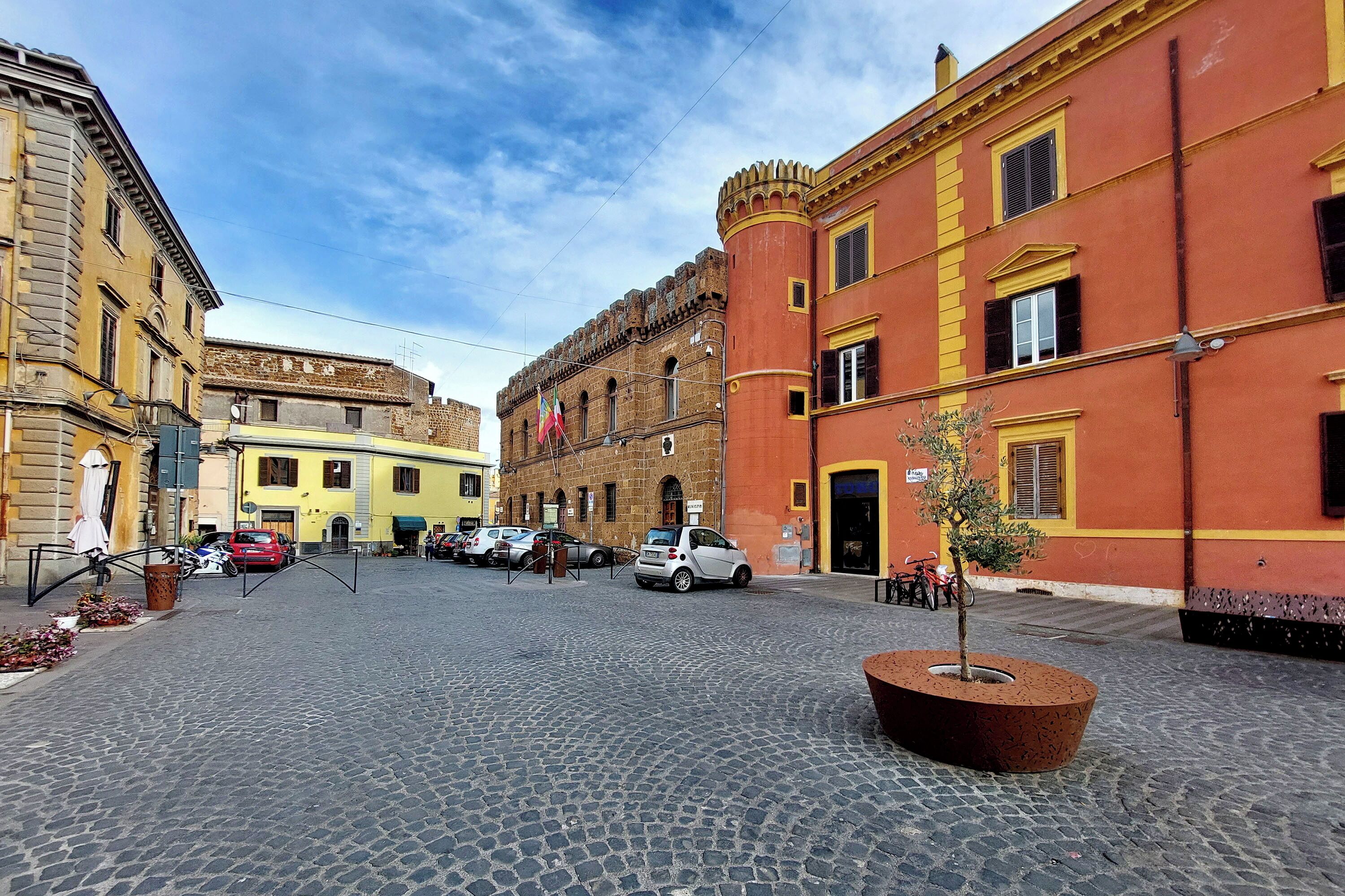 A cobblestone street with historic buildings, a small tree planter, and parked cars.