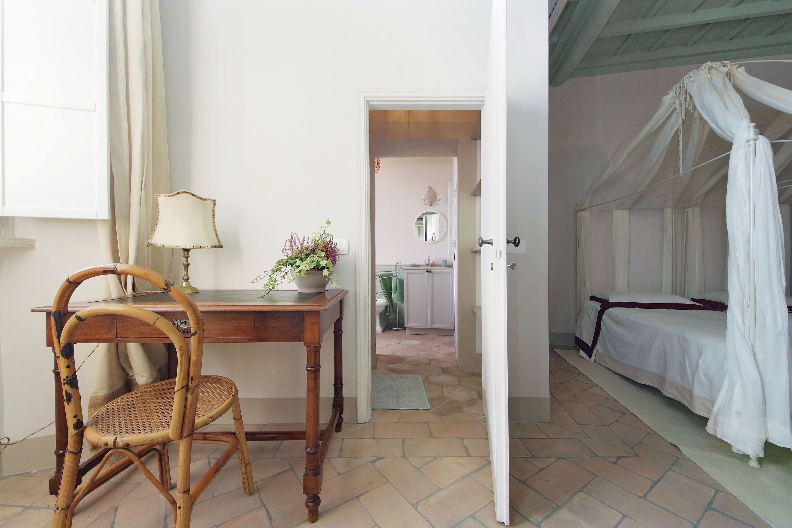 Una habitación con una cama, una silla, una mesa con una lámpara y una puerta que da a otra habitación.