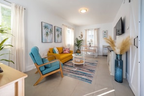 Ein Wohnzimmer mit gelbem Sofa, blauem Sessel, Couchtisch und an der Wand montiertem Fernseher.