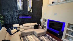 Un salon moderne avec un canapé d’angle, un téléviseur à écran plat, un mur décoré d’un motif et un ventilateur de plafond.