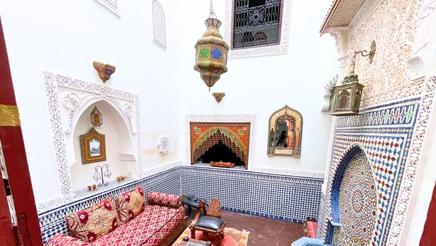 Ein traditionelles marokkanisches Interieur mit einer gemusterten Couch, einem kleinen Tisch, einer dekorativen Laterne und einer Kachelwand.