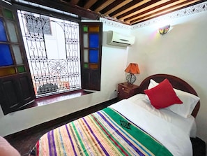 A hotel room with a bed, a nightstand with a lamp, an air conditioning unit, a window with decorative iron bars, and a patterned bedspread.