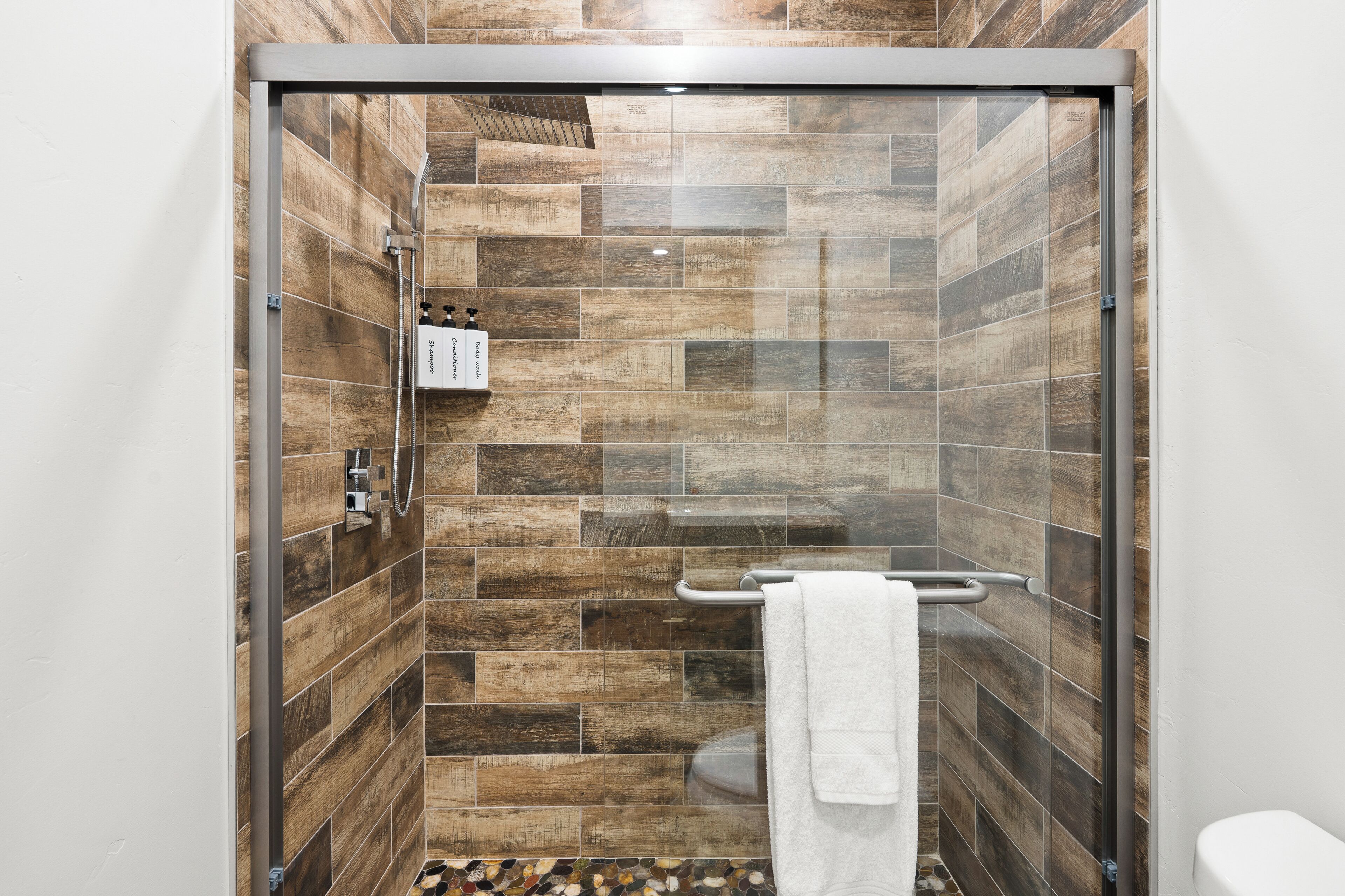 A shower with a glass door, wooden tiles, a towel rack, and a toilet visible to the right.