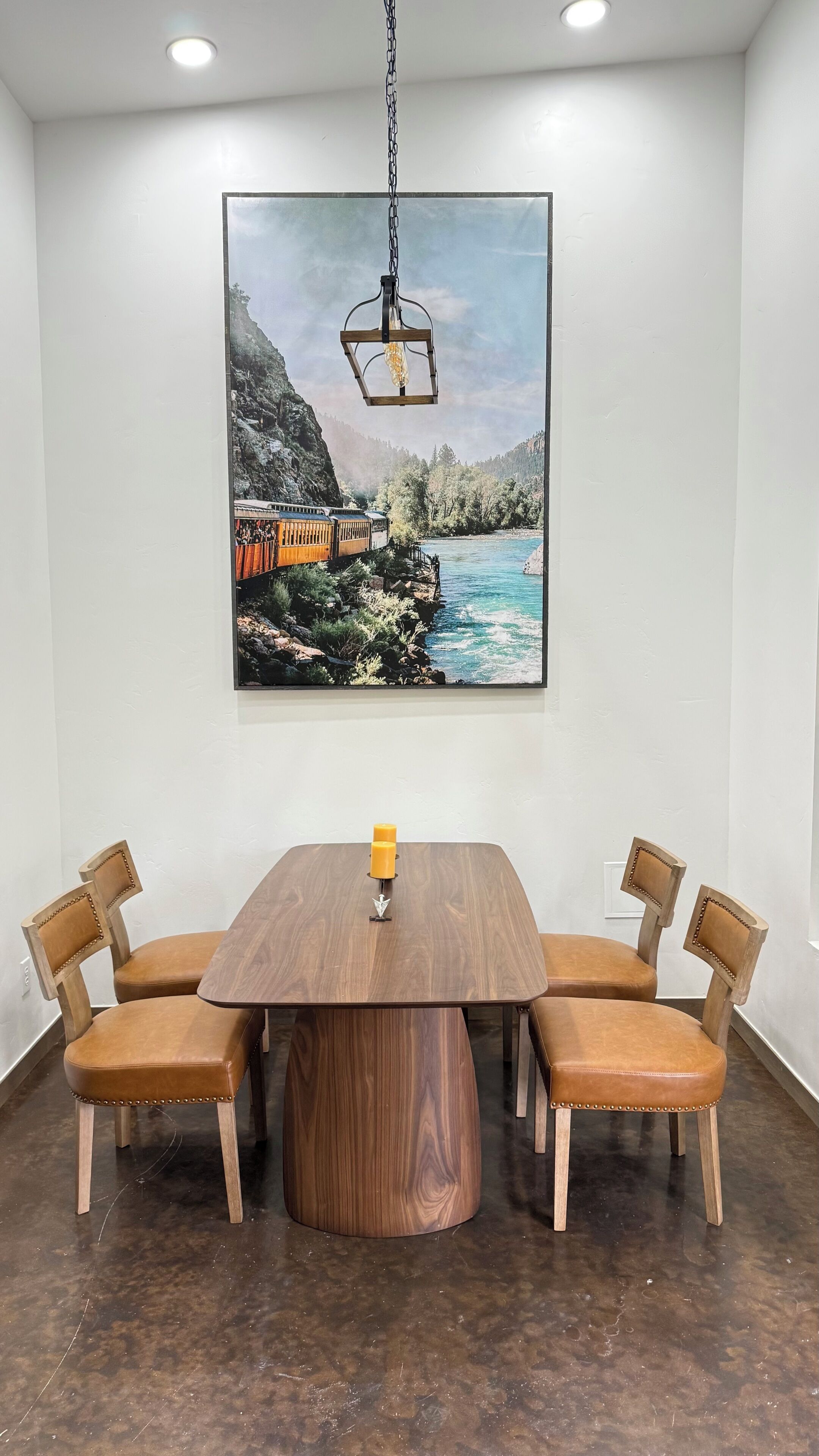 A dining area with a wooden table and chairs, a painting of a train on a cliff, and a hanging light fixture.