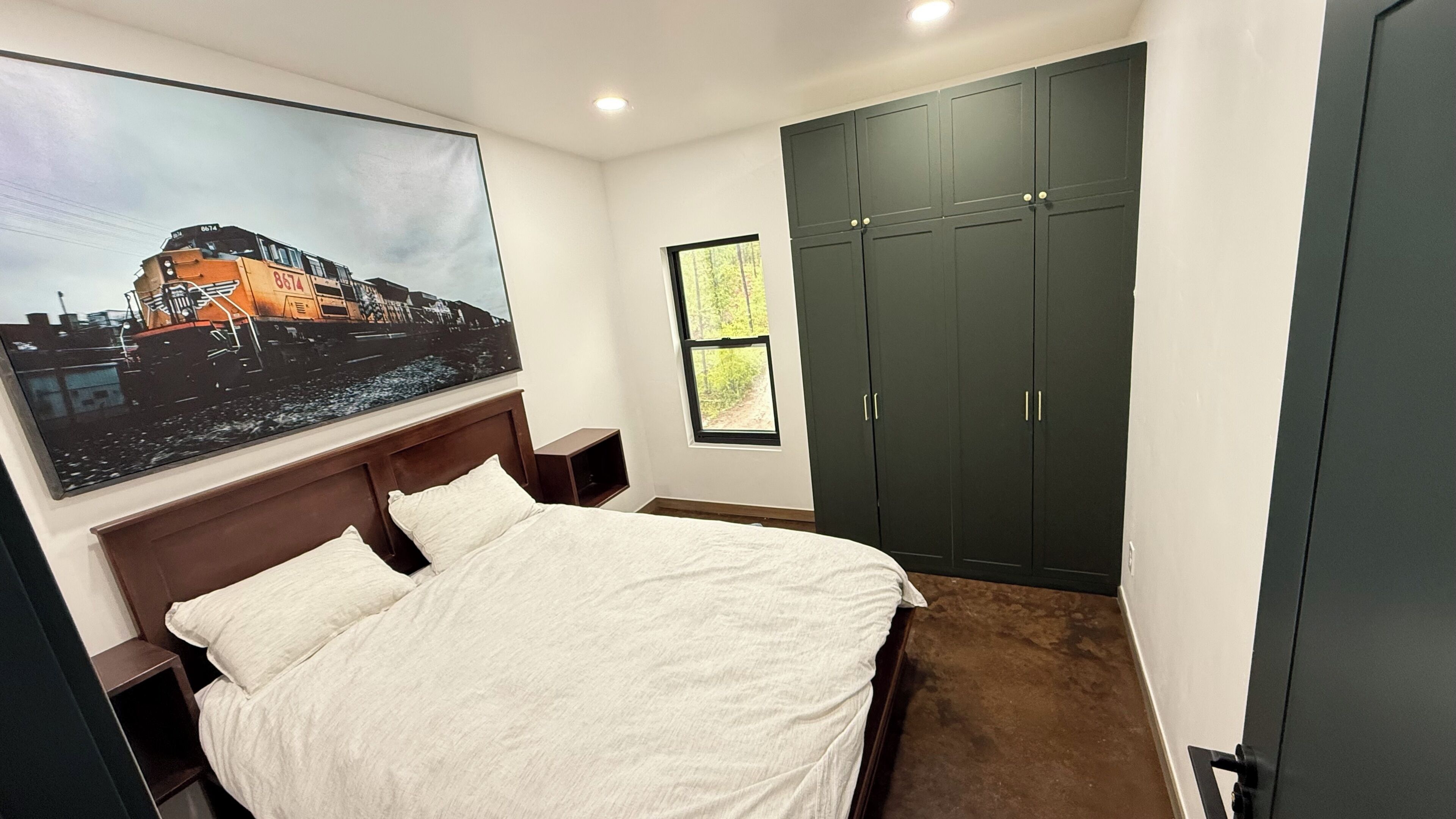 A bedroom with a bed, a large window, a wardrobe, and a painting on the wall.