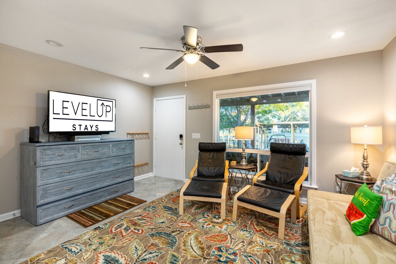A room with a ceiling fan, a TV on a dresser, two chairs, a sofa, a patterned rug, and a large window with a view.