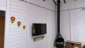 A room with white paneled walls, a TV mounted on the wall, a black chimney, a wooden chest, and a beige sofa. - Rancho Quemeninho - A refuge in the Atlantic Forest in the heart of PETAR. (Apiaí)