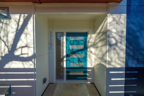 Ein Haus mit einer blauen Tür, der Hausnummer '7719C' an der Wand und dem Schatten eines Baumes.