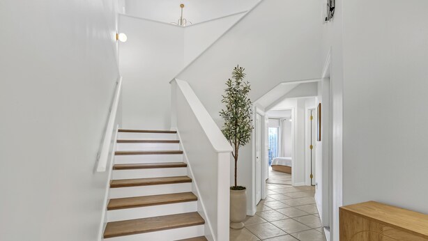 A staircase with wooden steps and white walls, leading to a hallway with a potted plant and a wooden bench. - Oceanside Vista - View of the Ocean from Balcony (Indialantic)