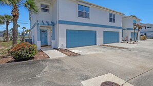 A two-story residential building with a blue garage door, a front door, and a small front yard with a palm tree and a bush. - Oceanside Vista - View of the Ocean from Balcony (Indialantic)