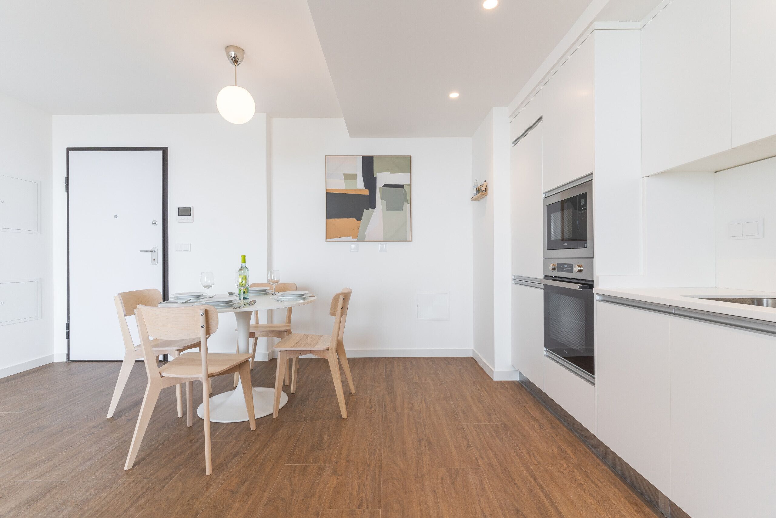 A modern kitchen with white cabinets, a built-in oven, and a dining area with wooden chairs and a round table.