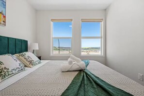 Ein Schlafzimmer mit Bett, Kissen, Decke, Nachttisch, Lampe und zwei Fenstern mit Landschaftsblick.