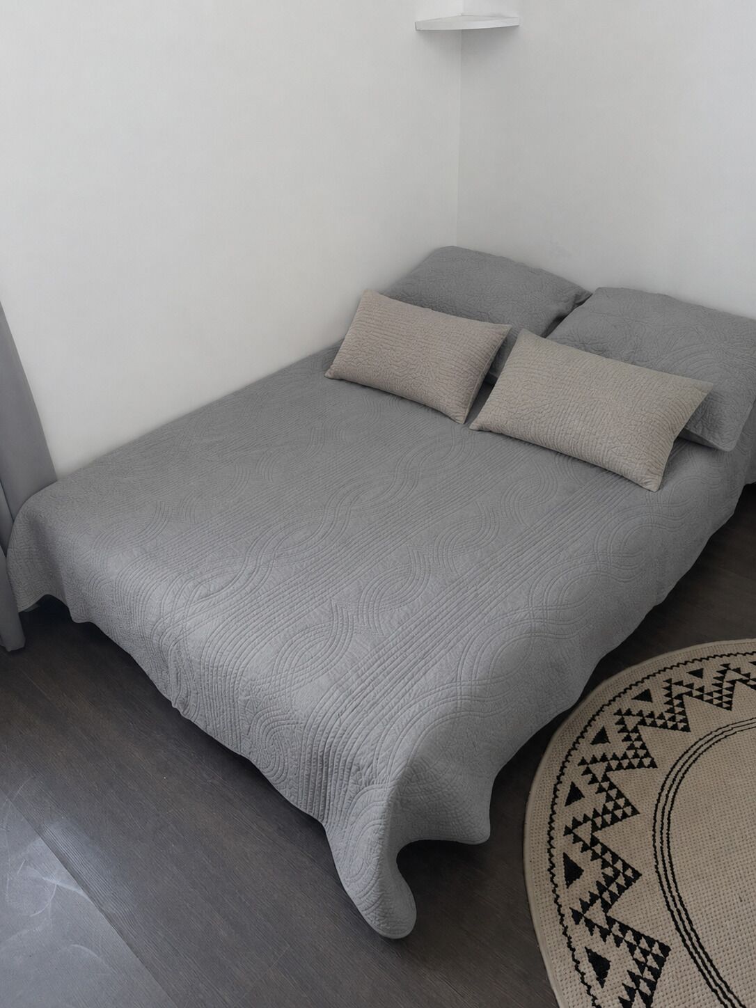 Ein Einzelbett mit einer strukturierten, grauen Bettdecke und Kissen, ein runder Teppich mit geometrischem Muster und eine weiße Wand.