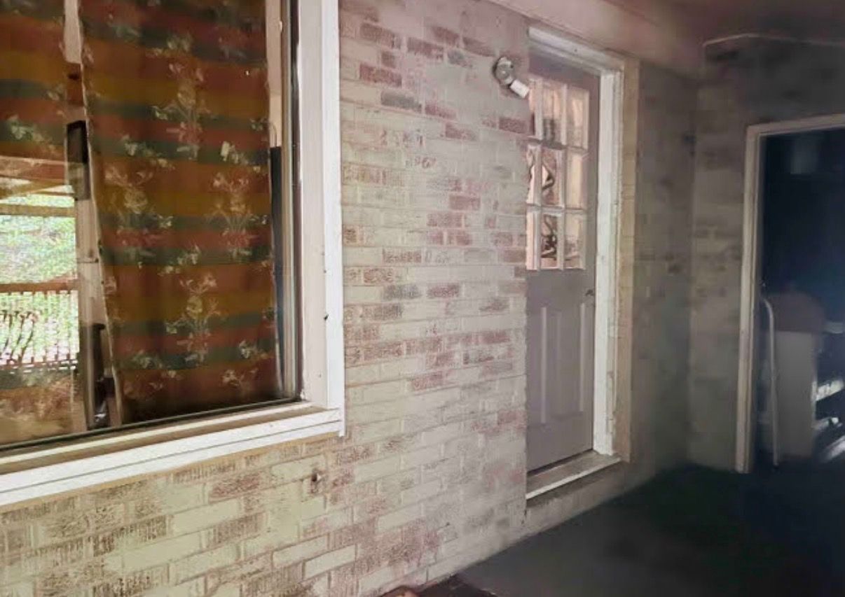 A room with a brick wall, a window with a floral curtain, and a door with a small shelf.