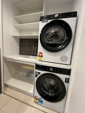 A laundry room with two front-loading washing machines, a shelf above, and a tiled backsplash. - Riverfront | Balcony | Southbank Promenade | Arts Precinct | Parking | Crown (Southbank)