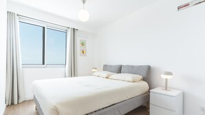 A bedroom with a bed, bedside tables, a nightstand, a window, and a wall-mounted light fixture. - Infinity Blue by Atlantic Holiday (Camara de Lobos)