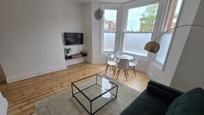 Ein modernes Wohnzimmer mit einem Glastisch, einem grünen Sofa, einem Fernsehgerät an der Wand, einem Essbereich mit Stühlen und großen Fenstern.