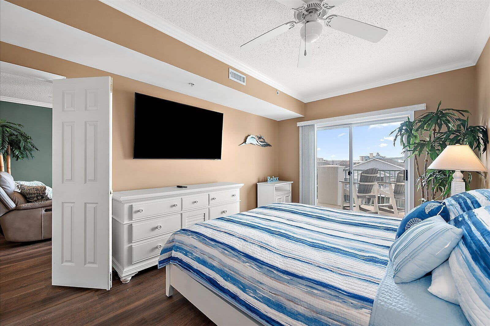 A bedroom with a bed, a TV, a dresser, a desk, a ceiling fan, and a view of buildings through a window.