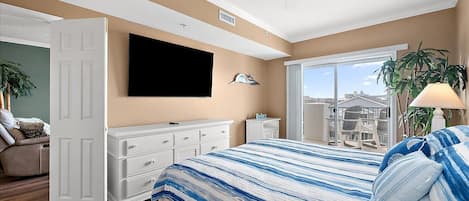 A bedroom with a bed, a TV, a dresser, a desk, a ceiling fan, and a view of buildings through a window.