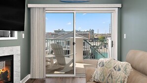 A living room with a fireplace, a large sliding glass door leading to a balcony, a sign reading 'Beach House,' and a view of buildings and a marina. - Three Bedroom Oceanside Condo with Great Amenities (Ocean City)
