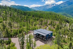 Une cabane surmontée d’un toit en métal, entourée d’une forêt dense et avec des montagnes en arrière-plan.