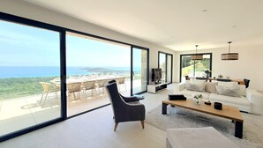 A modern living room with a large sliding glass door leading to an outdoor dining area, a sofa, a coffee table, and a TV. - Villa Sarra Di Pinarellu (Zonza)