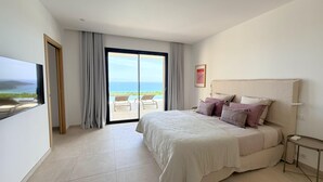 A bedroom with a bed, a TV, a large window with curtains, and a view of the sea. - Villa Sarra Di Pinarellu (Zonza)