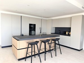 A modern kitchen with a central island, bar stools, and built-in appliances. - Villa Sarra Di Pinarellu (Zonza)