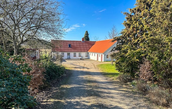 Ein ländliches Anwesen mit einem Schotterweg, der zu einem weißen Gebäude mit rotem Dach führt, umgeben von Bäumen und Sträuchern.