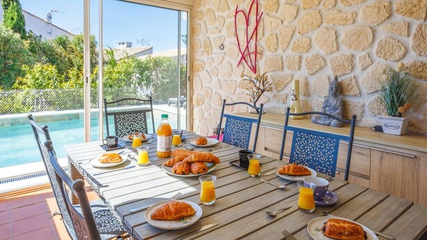 A dining table with breakfast items, a pool, and a stone wall with decorative elements. - Villa Repose by Villa Plus (Argeliers)