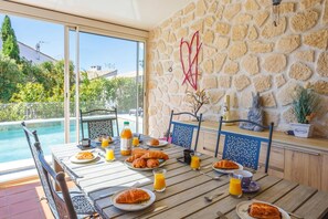 A dining table with breakfast items, a pool, and a stone wall with decorative elements. - Villa Repose by Villa Plus (Argeliers)