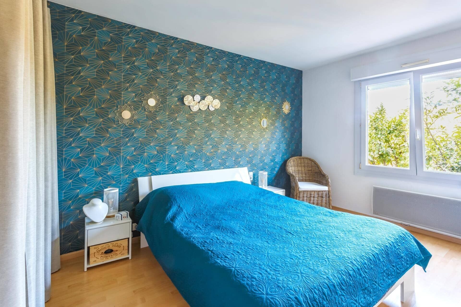 Ein Schlafzimmer mit einer Wand mit geometrischem Muster, einem Bett mit blauer Bettdecke, einem Korbsessel, einem Nachttisch mit einer Flasche und einem Fenster mit Blick auf Bäume.