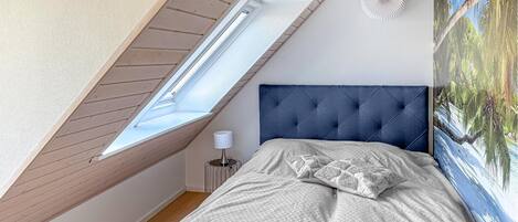 Un dormitorio con techo inclinado, una cama con cabecera capitonada, una mesita de noche, una lámpara y un aplique de pared.