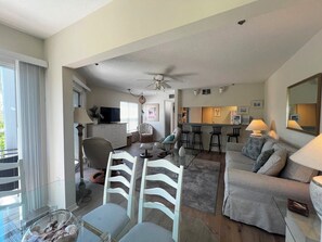 Ein modernes Wohnzimmer mit einem Glastisch, weißen Stühlen, einem grauen Sofa, einer Küchenzeile mit Barhocker und einem Deckenventilator.