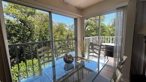 Una stanza con tavolo in vetro, sedie, ventilatore a soffitto e vista su alberi e un balcone.