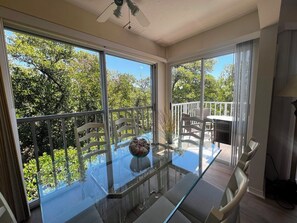 Ein Zimmer mit Glastisch, Stühlen, Deckenventilator und Blick auf Bäume sowie einen Balkon.