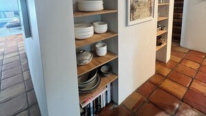 Een hoek van een kamer met een wandplank waarop diverse serviesdelen en een lijst met een kunstwerk staan.