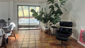 Een woonkamer met een grote rubberplant, een zwarte leren fauteuil, een witte bank, een glazen salontafel en een schuifpui die leidt naar een balkon.