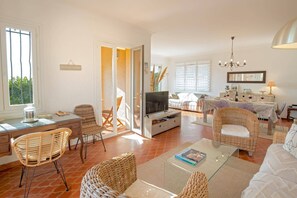 Ein geräumiges Wohnzimmer mit Fernseher, Korbmöbeln, einem Essbereich mit Tisch und Stühlen sowie einer Kandelaber.