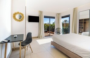Ein modernes Hotelzimmer mit Bett, Schreibtisch, Stuhl, Fernseher und einem Balkon mit Blick auf Palmen.