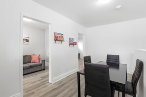Una sala de estar moderna con un sofá gris, una mesa de comedor con cuatro sillas y un pasillo que conduce a otra habitación.