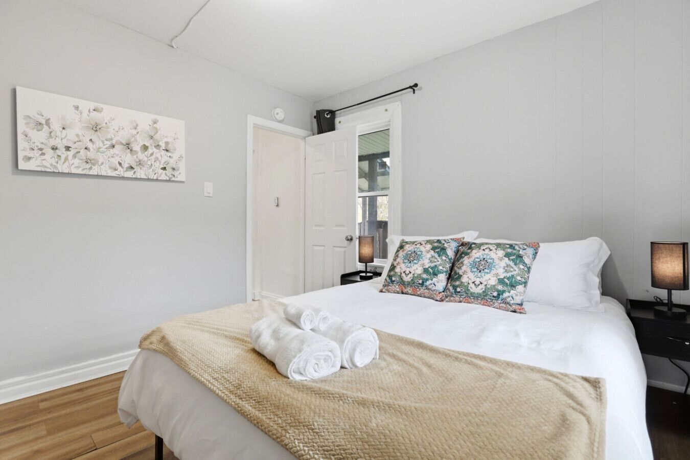 A bedroom with a bed, pillows, a bedside table, a lamp, a door, and a wall-mounted artwork.