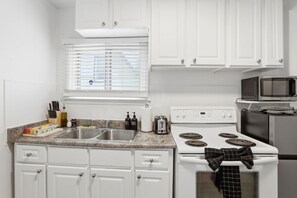 A kitchen with white cabinets, a double sink, a stove, a microwave, and a window with blinds.